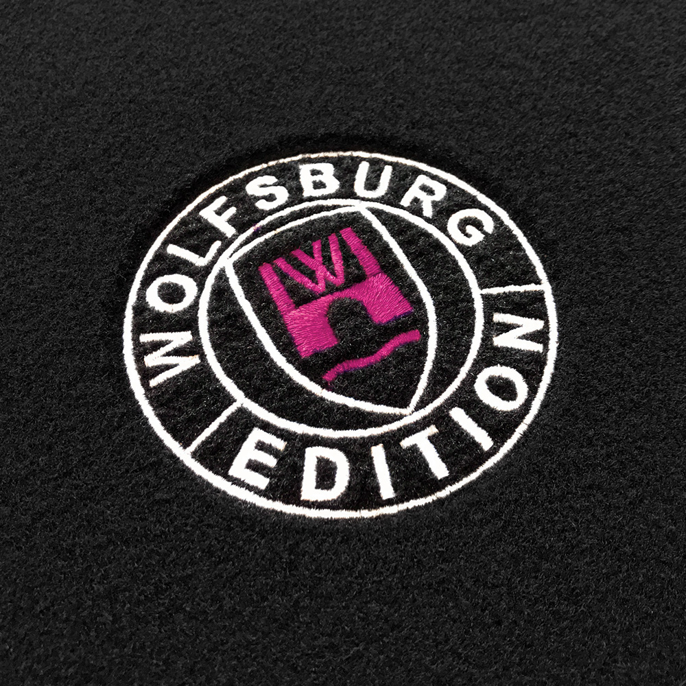 Das gestickte Wolfsburg Editions Logo auf einem schwarzen Teppich und das Logo ist in pink.